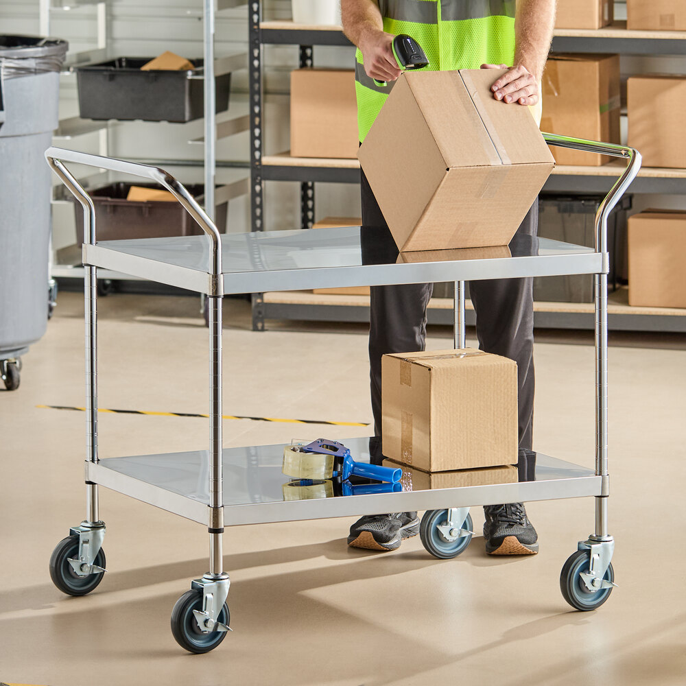 A two-shelf stainless steel utility cart with four swivel casters, holding cardboard boxes and packing tape, in a warehouse setting.