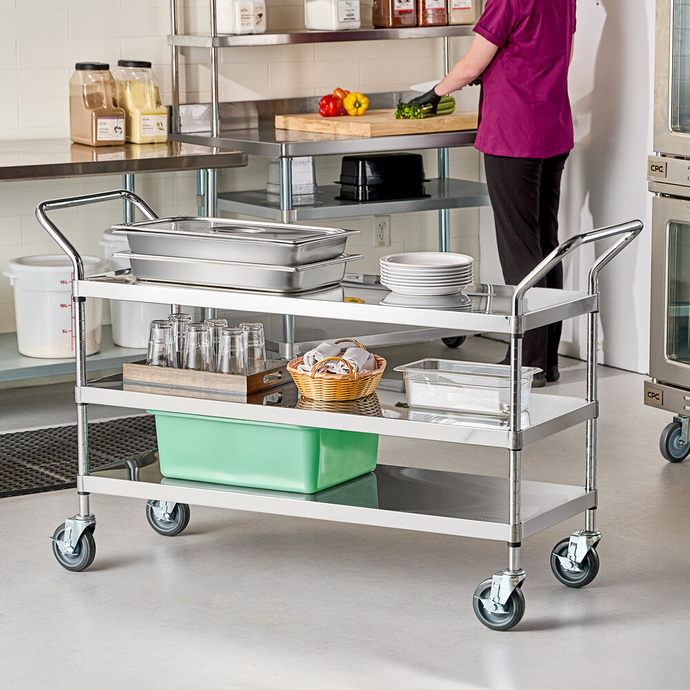 A stainless steel utility cart with three shelves, holding trays, plates, glasses, a basket, and a green bin, situated in a commercial kitchen setting.