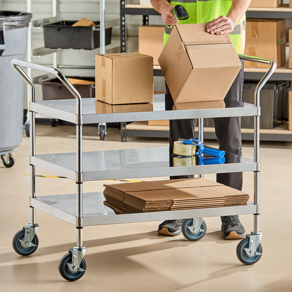 A three-shelf stainless steel utility cart with handles and caster wheels, holding cardboard boxes, packing tape, and flat boxes, with a person working nearby.