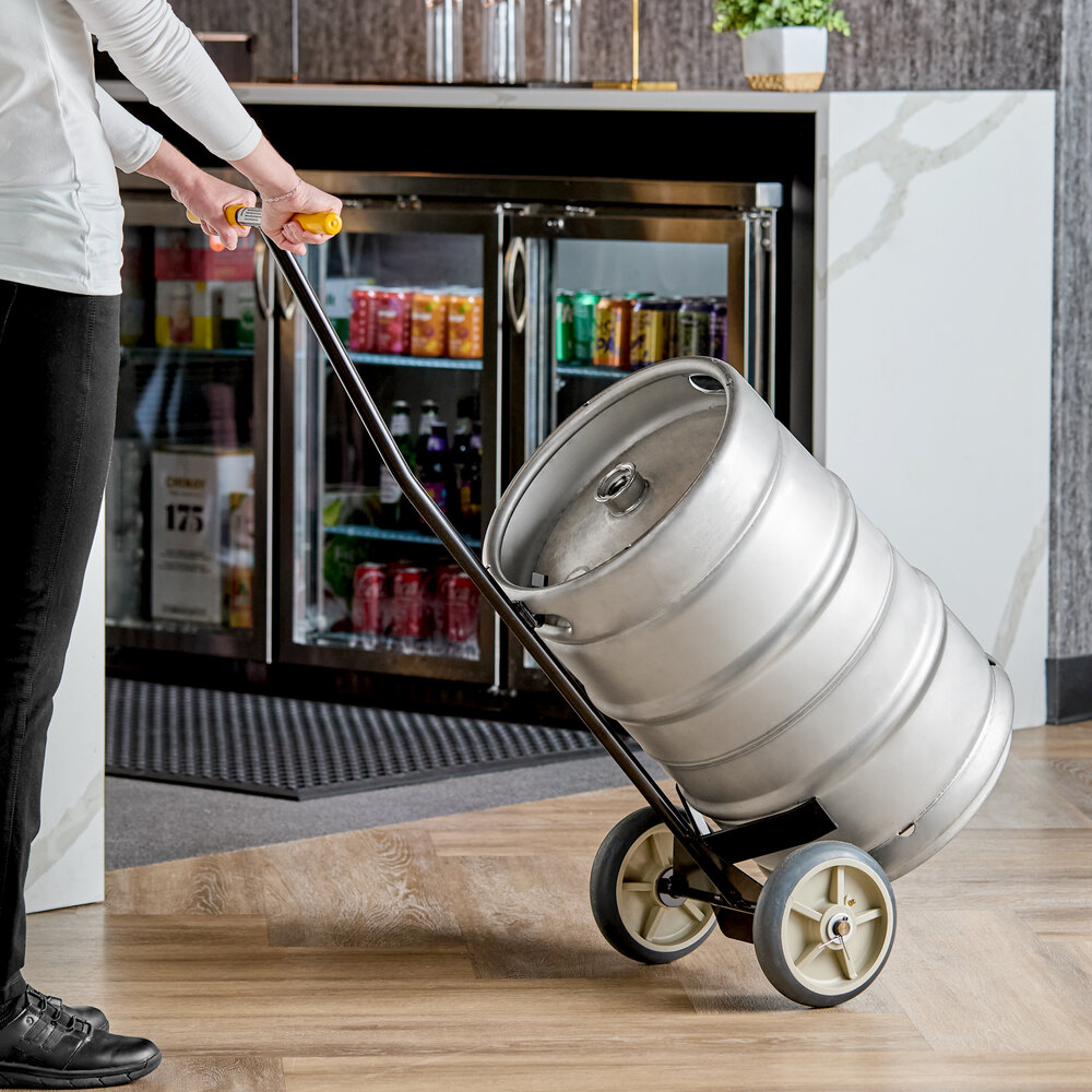 A person is using a hand truck to transport a large metal keg indoors.