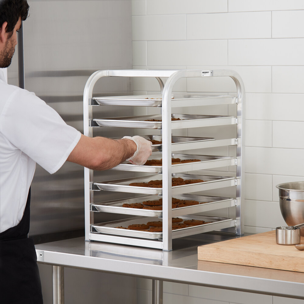 A person is placing trays of cookies onto a multi-tiered metal rack on a stainless steel countertop in a kitchen setting.