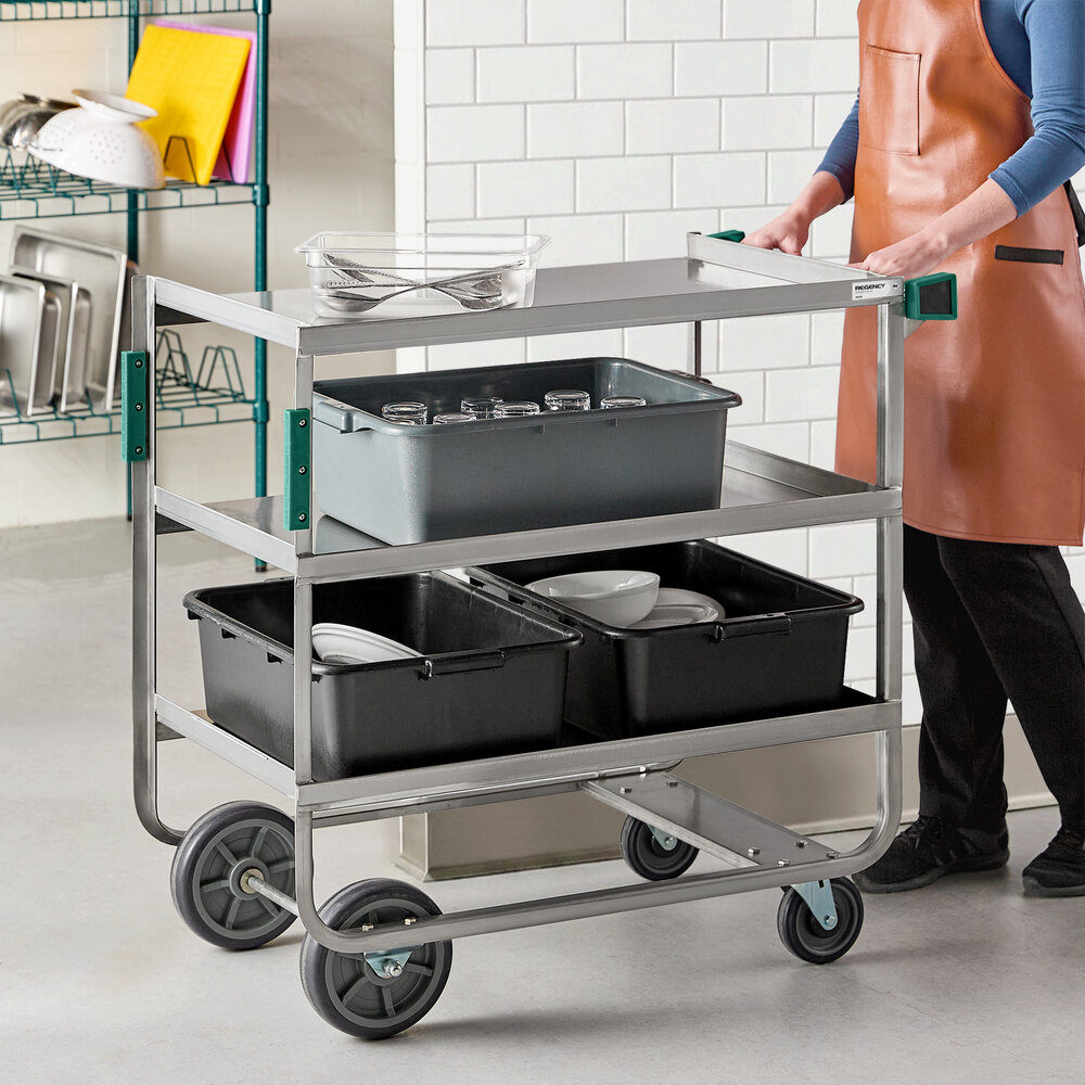 A person is pushing a three-shelf stainless steel cart loaded with bins and kitchenware in a commercial kitchen setting.