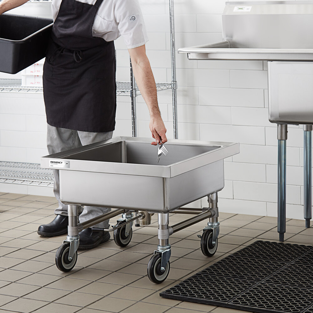 A person in a kitchen is placing utensils into a large, square, stainless steel soaking sink on wheels.