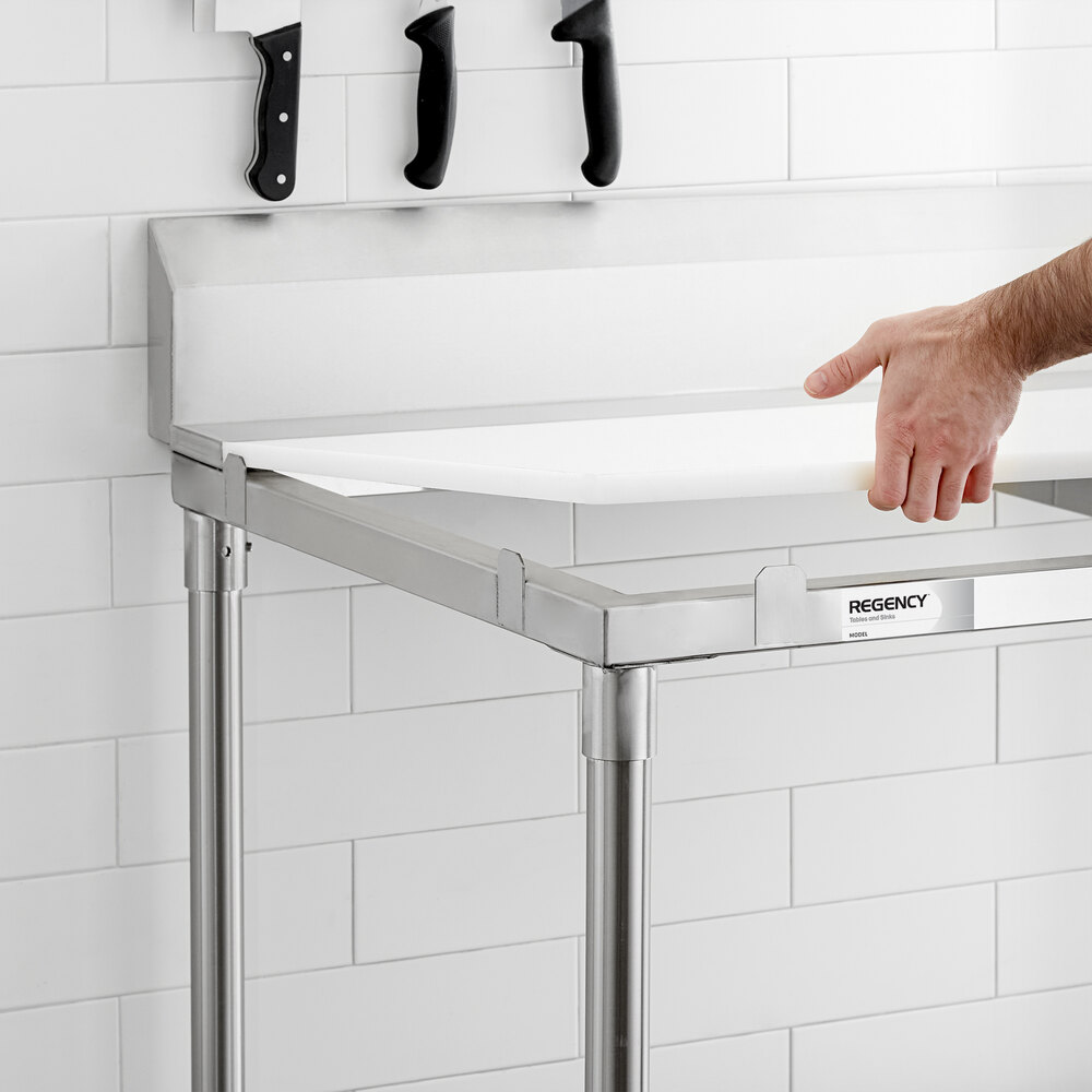 A person is placing a white tabletop onto a stainless steel table frame in a kitchen with white tiled walls and knives hanging on a magnetic strip above.