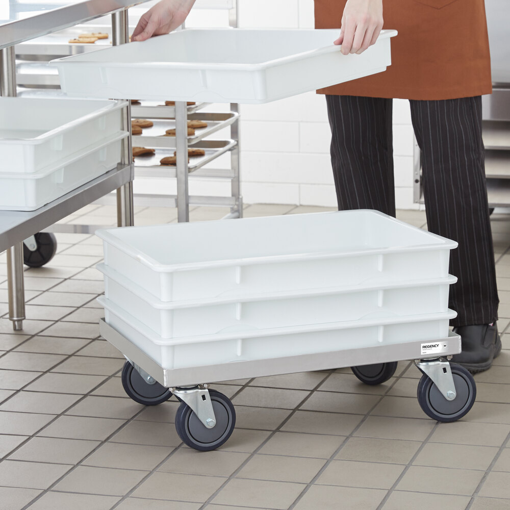 A person is stacking large white rectangular trays on a metal cart with four wheels in a commercial kitchen setting.
