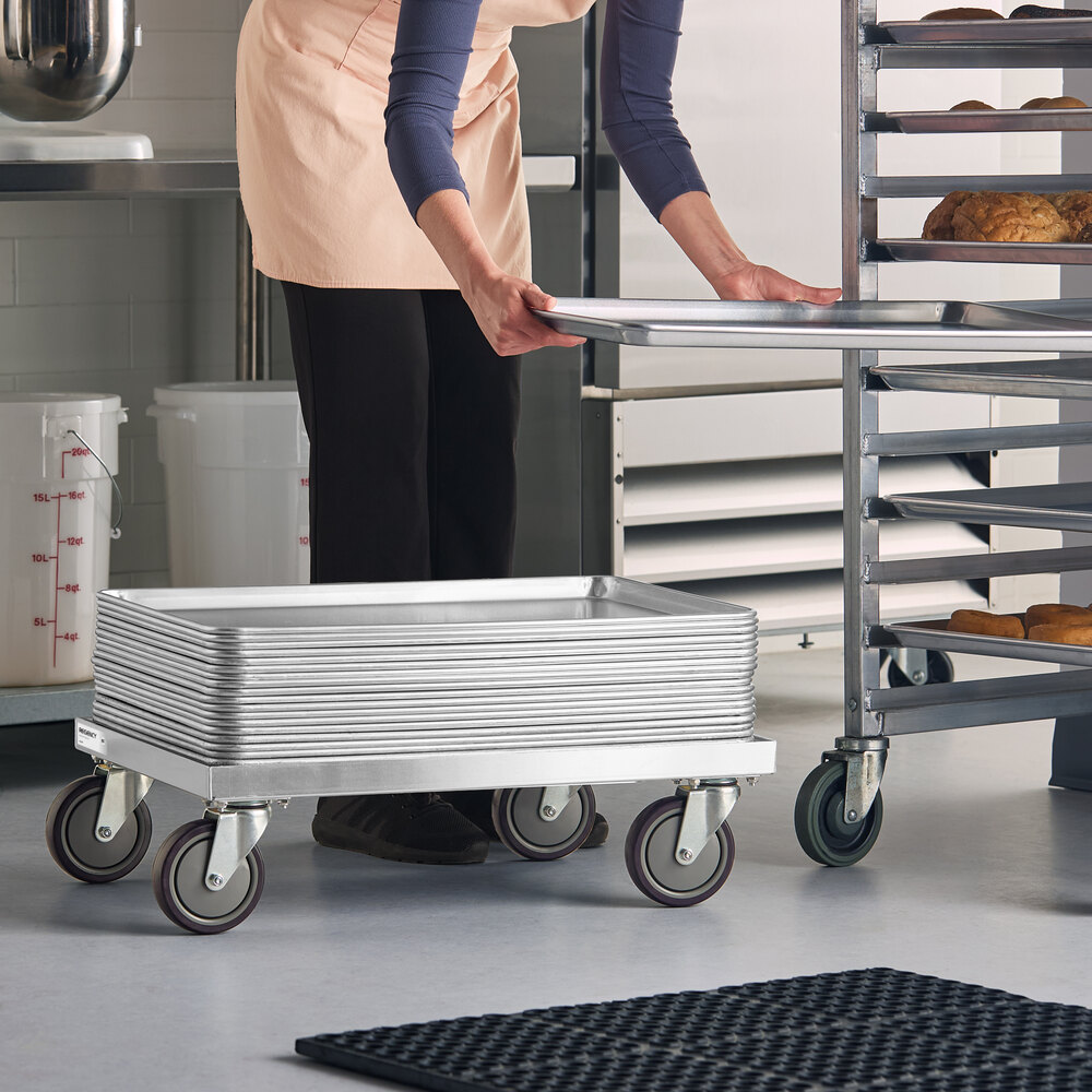 A stack of metal trays is placed on a low, wheeled dolly in a commercial kitchen setting, with a person transferring a tray to a nearby rack.