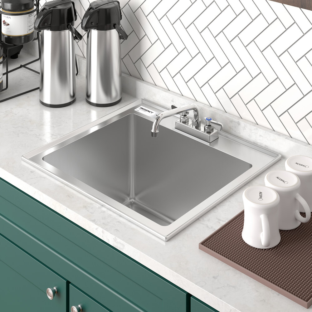 A stainless steel single-basin sink with a faucet, set in a white countertop with green cabinetry and surrounded by coffee dispensers and mugs.