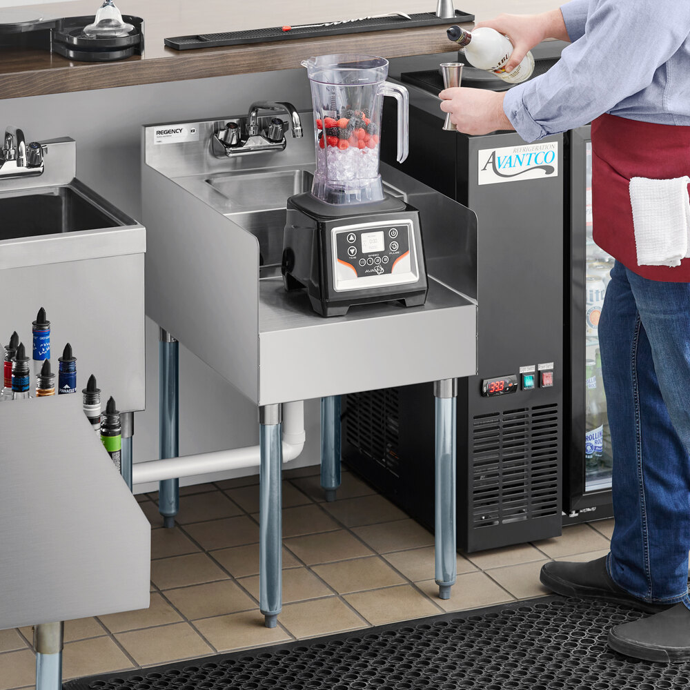 A stainless steel blender station with a dump sink, featuring a blender with a pitcher containing ice and berries, and a person preparing to pour liquid into a measuring cup.