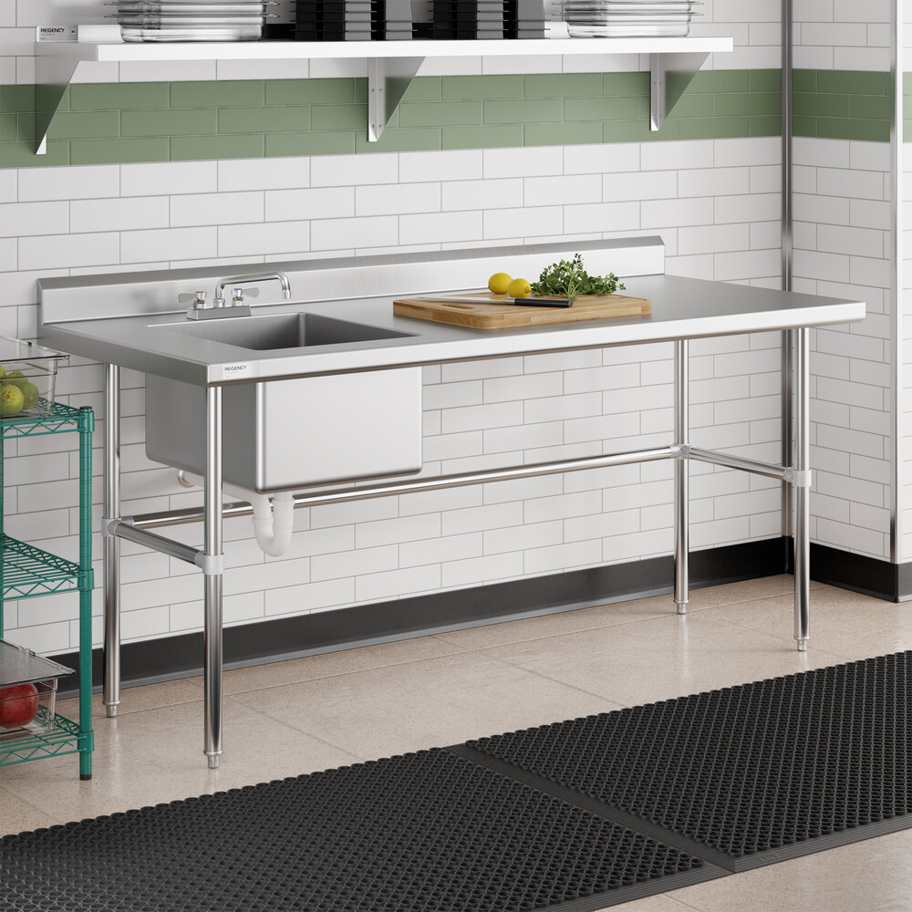 A stainless steel work table with a left-side sink, faucet, and cross bracing, situated in a tiled kitchen area with a cutting board and vegetables on top.