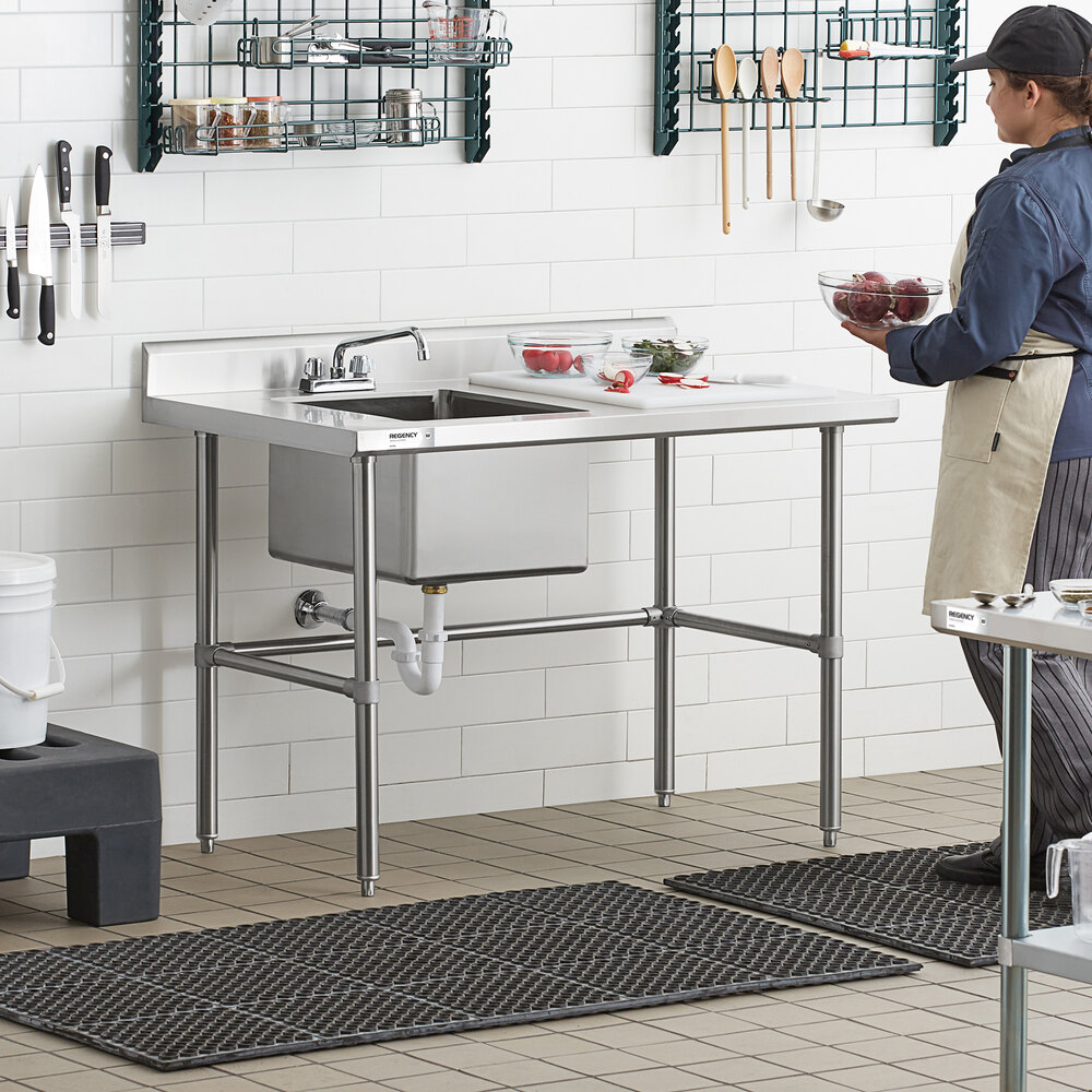 A stainless steel work table with a sink, cross brace support, and a person preparing food beside it in a commercial kitchen setting.