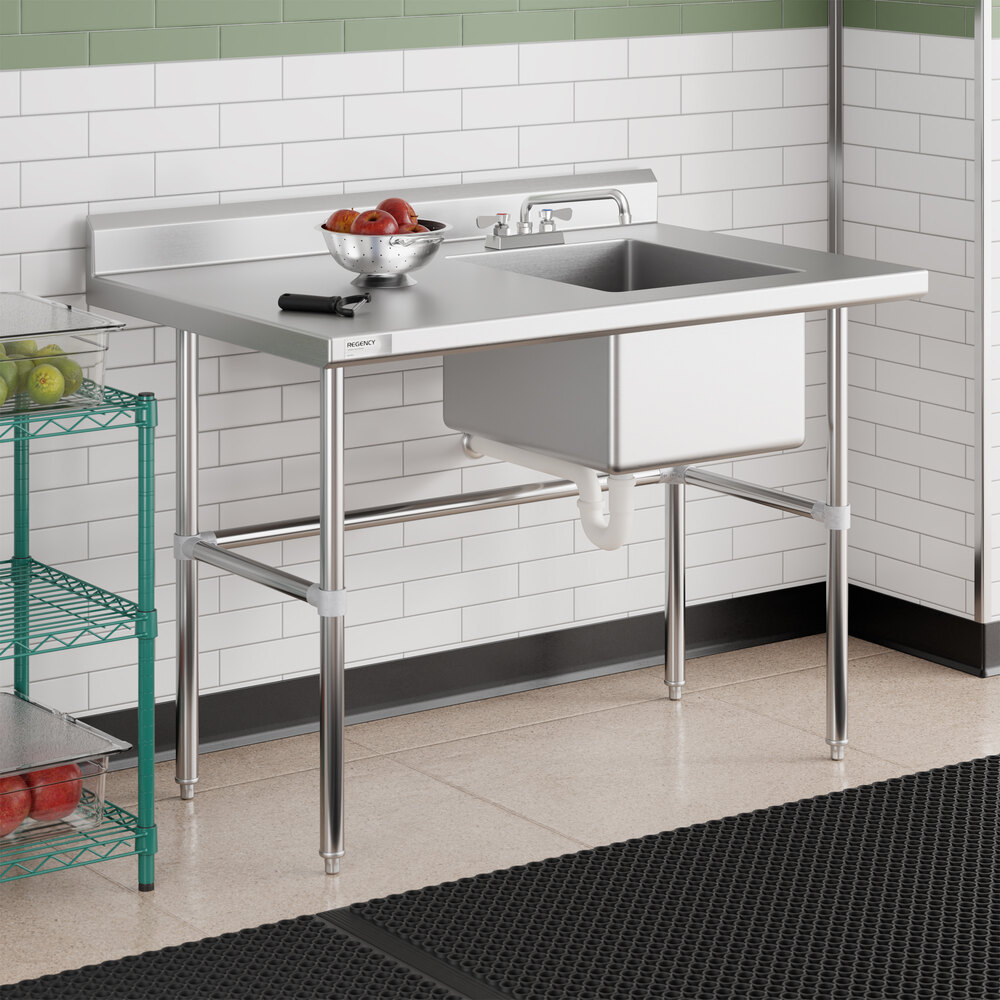 A stainless steel work table with a right-side sink, faucet, and cross bracing, situated in a tiled room with a bowl of fruit and a utensil on top.