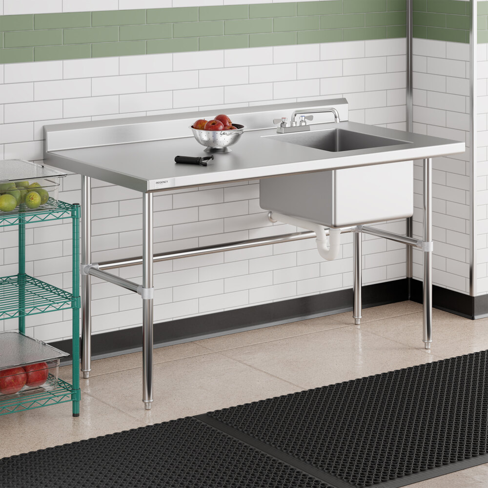 A stainless steel work table with a right-side sink, faucet, and cross bracing, placed against a tiled wall with a bowl of fruit and a knife on top.