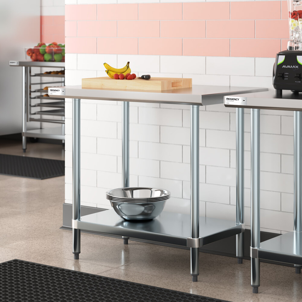 A stainless steel work table with a lower shelf, holding a metal mixing bowl underneath and a cutting board with fruit and a knife on top.