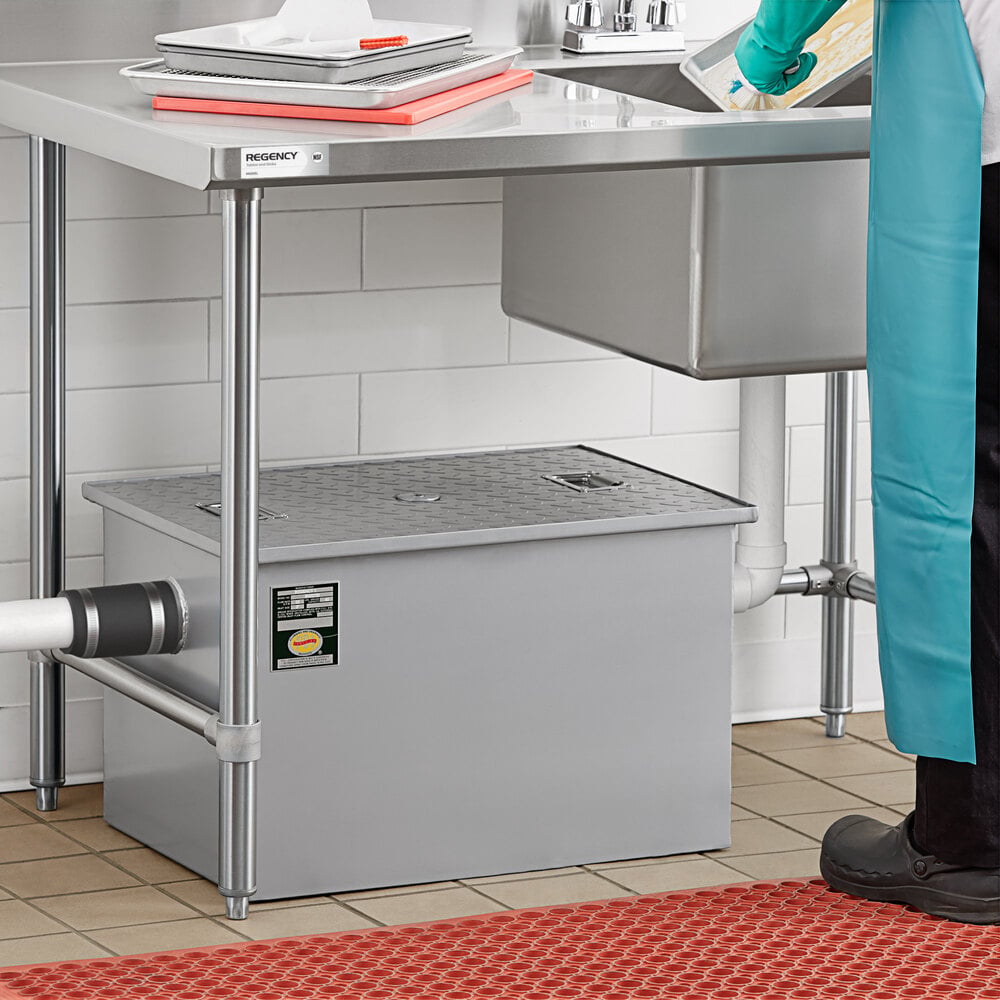 A large rectangular metal box with a textured lid and pipe connections is positioned under a commercial sink in a tiled kitchen area.
