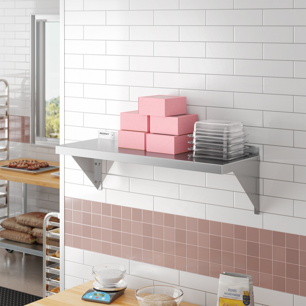 A stainless steel wall-mounted shelf holding pink boxes and clear trays in a commercial kitchen setting.