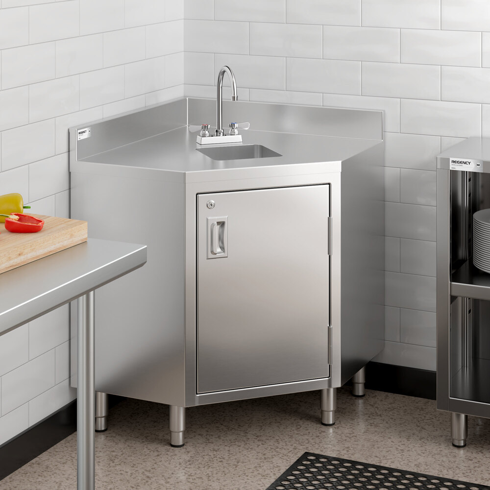 A stainless steel corner work table with an integrated sink, faucet, and a hinged door cabinet below, situated in a tiled kitchen corner.