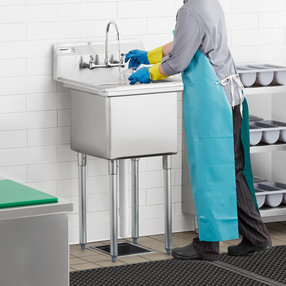 A person wearing gloves and a blue apron is using a stainless steel commercial sink with four metal legs in a tiled room.