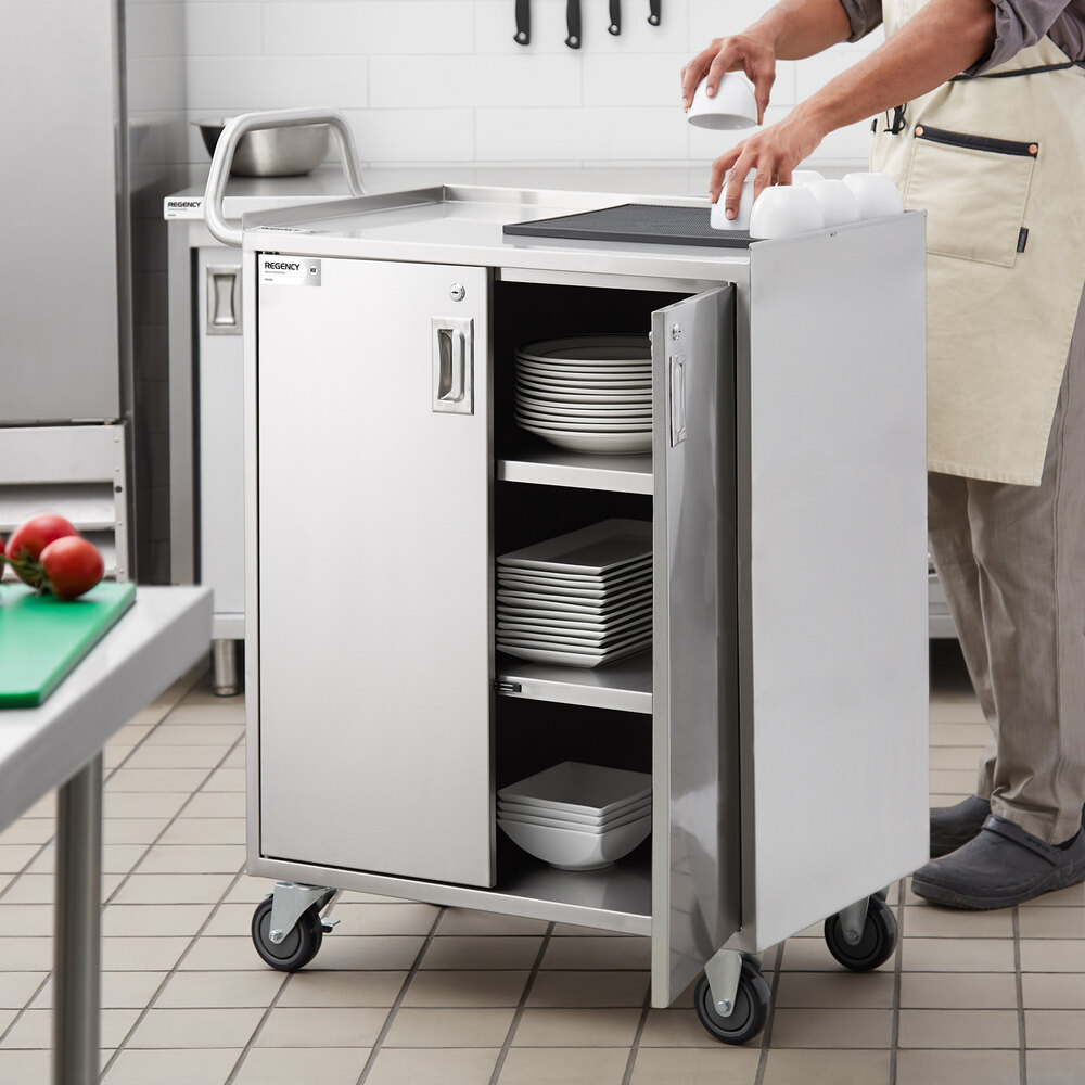 A stainless steel utility cart with wheels, an enclosed base, locking doors, and shelves holding plates and bowls, situated in a commercial kitchen.