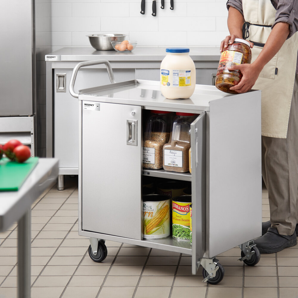 A stainless steel utility cart with three shelves, locking doors, and wheels, shown in a kitchen setting with various food containers stored inside and on top.