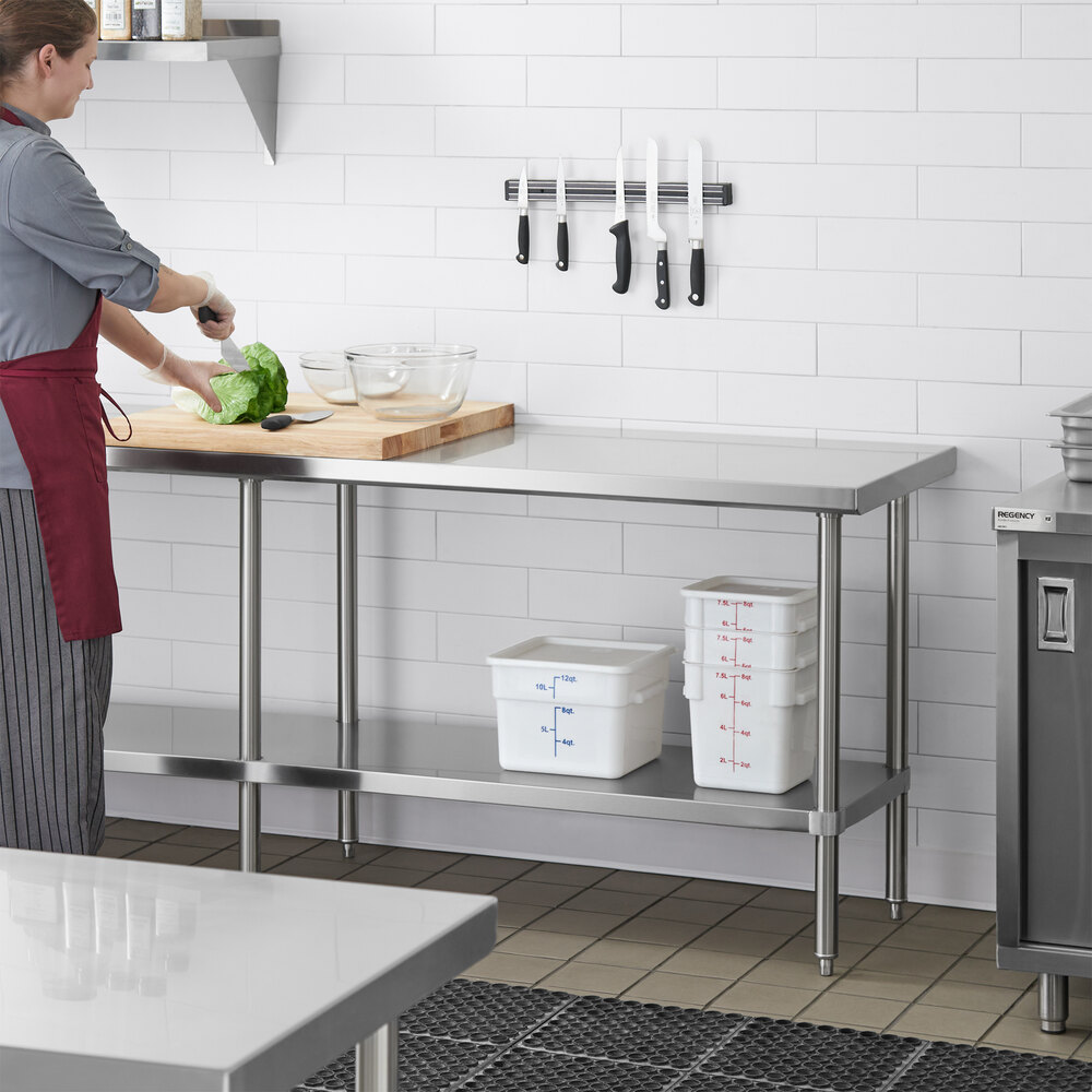 A person is preparing food on a stainless steel work table with an undershelf, which holds several plastic storage containers; knives are mounted on the wall above the table.