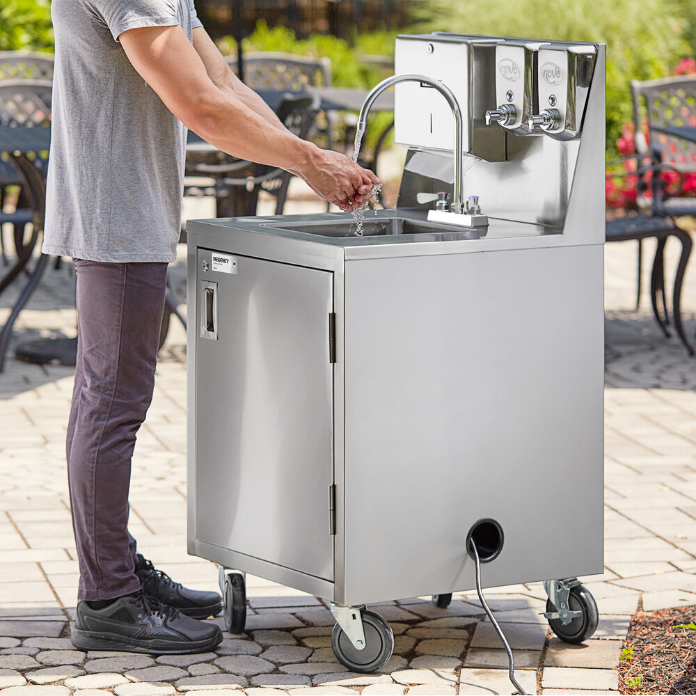 A person is washing their hands at a stainless steel portable hand sink cart with wheels, a faucet, and soap dispensers, set outdoors on a paved surface.