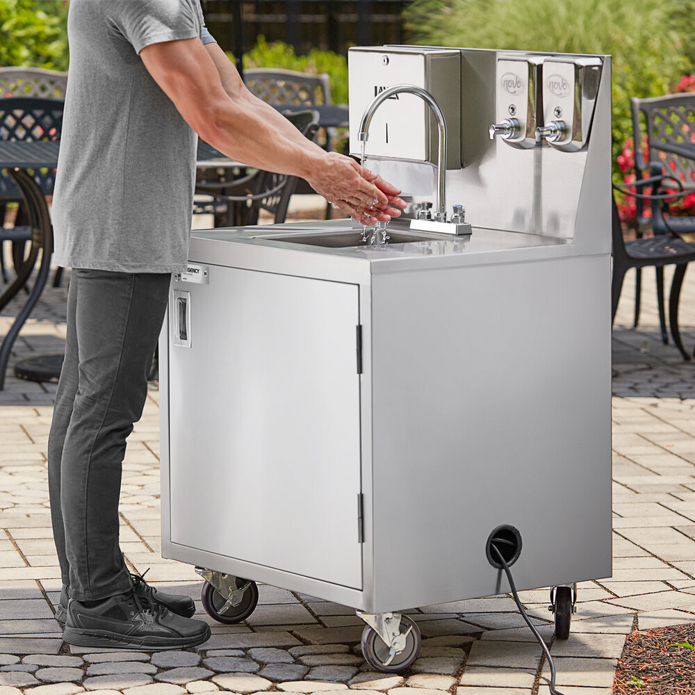 A person is washing their hands at a stainless steel portable hand sink cart with wheels, a faucet, and soap dispensers, set outdoors on a paved surface.