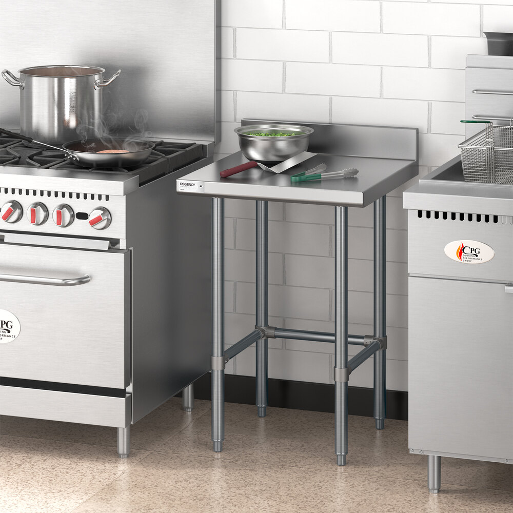 A stainless steel work table with an open base and a 4-inch backsplash, positioned between a stove and a fryer in a commercial kitchen, with a metal bowl and utensils on top.