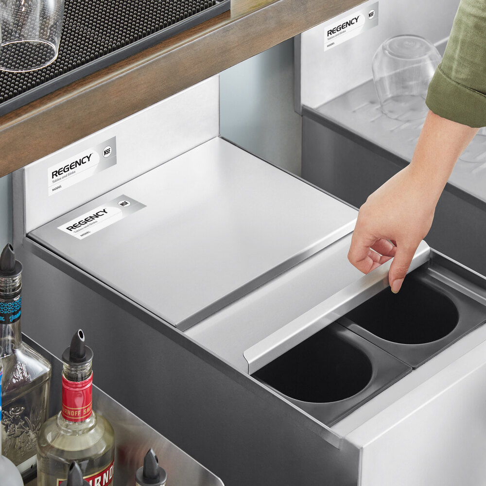 A person is sliding open a stainless steel lid on a rectangular underbar ice bin, with bottles and glasses nearby.
