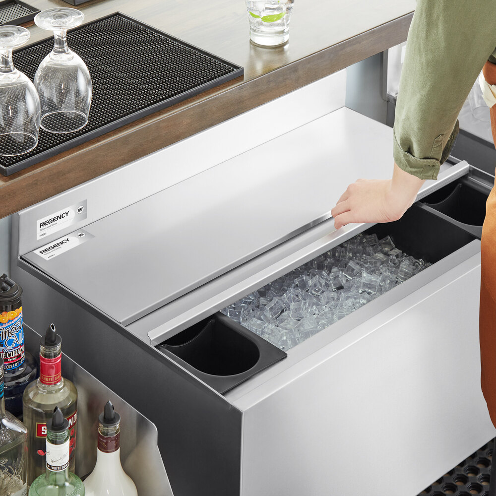 A person is sliding open the stainless steel lid of an underbar ice bin filled with ice, with bottles and glassware nearby.