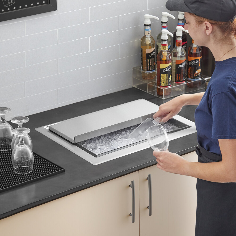 A person is scooping ice from a stainless steel ice bin with a sliding lid that is installed in a countertop.