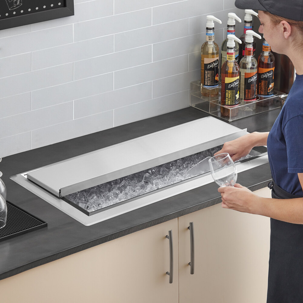 A person is scooping ice from a large stainless steel ice bin with a sliding lid, installed in a countertop.