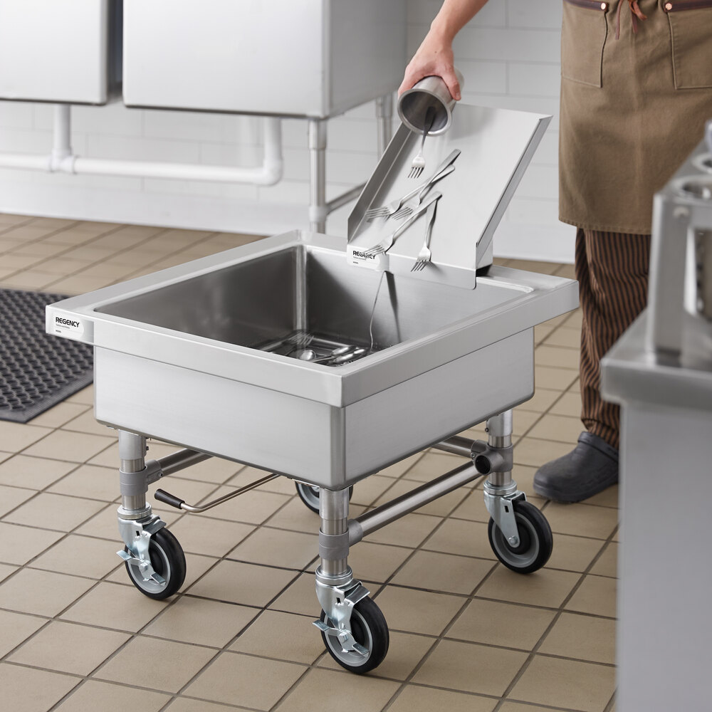 A person is pouring silverware and water from a detachable chute into a stainless steel mobile soaking sink on wheels in a commercial kitchen setting.