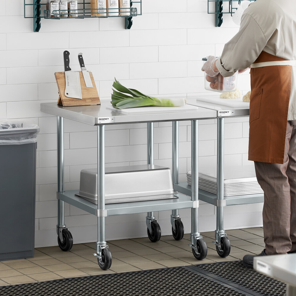 A stainless steel work table with a lower shelf and casters, shown in a kitchen setting with cutting boards, knives, and vegetables on top.