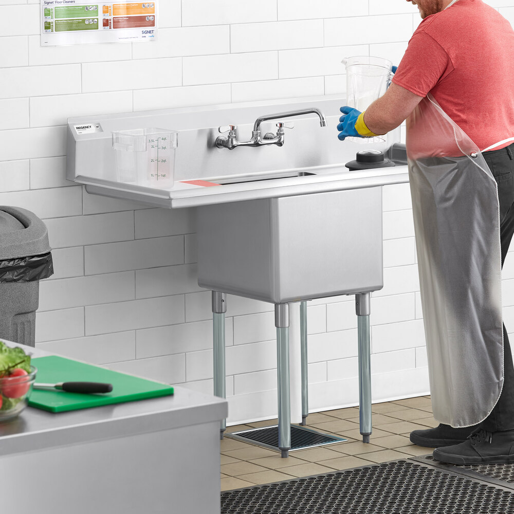 A person wearing gloves and an apron is working at a stainless steel sink with two drainboards and a deep sink bowl, set against a tiled wall in a commercial kitchen environment.