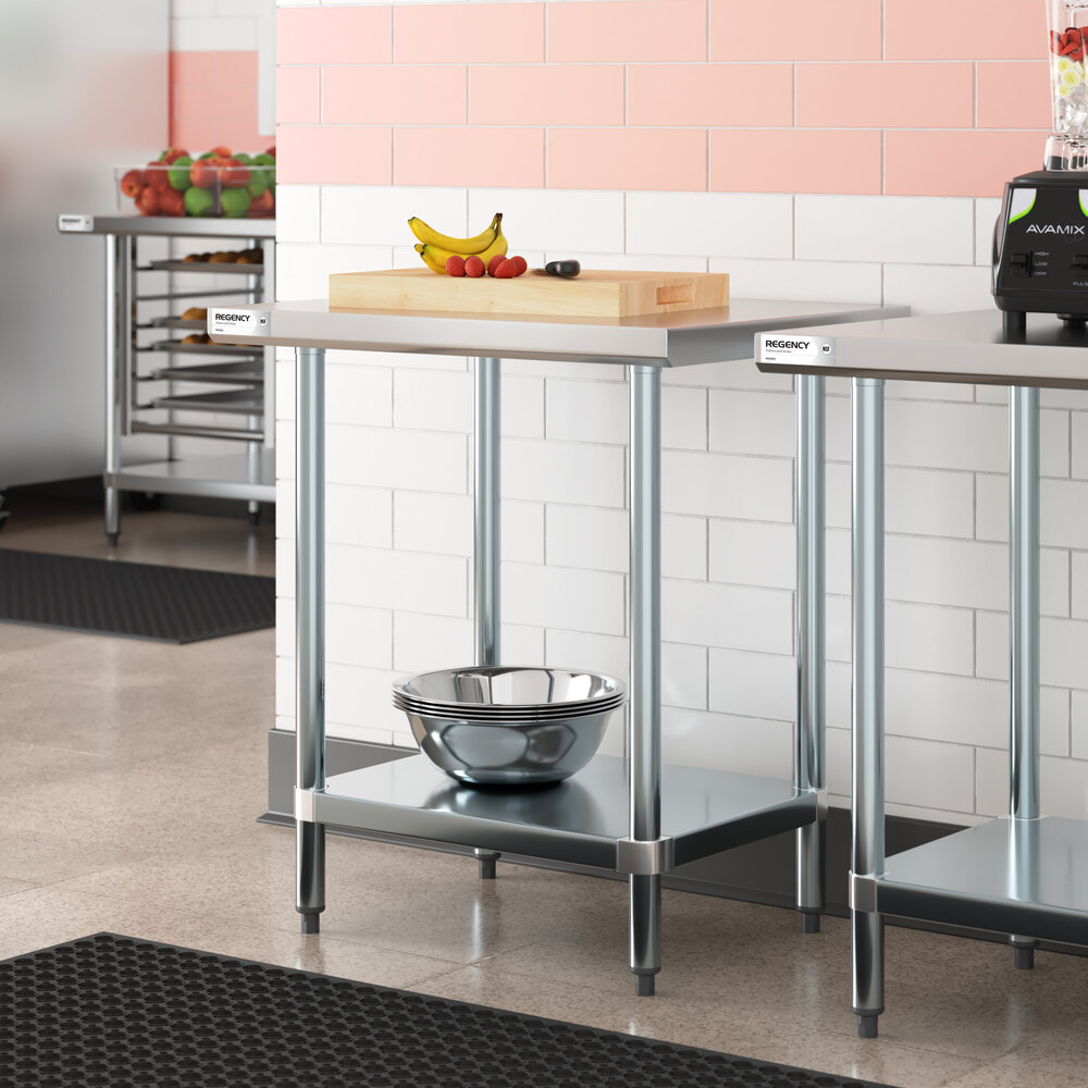 A stainless steel work table with a lower shelf, holding a metal mixing bowl, and a wooden cutting board with fruit on top.