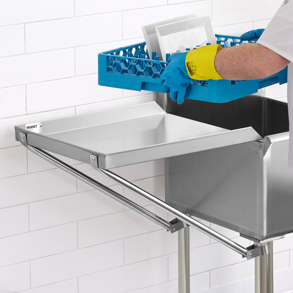A person wearing blue and yellow gloves is placing a blue dish rack with plates onto a stainless steel detachable drainboard attached to a sink.