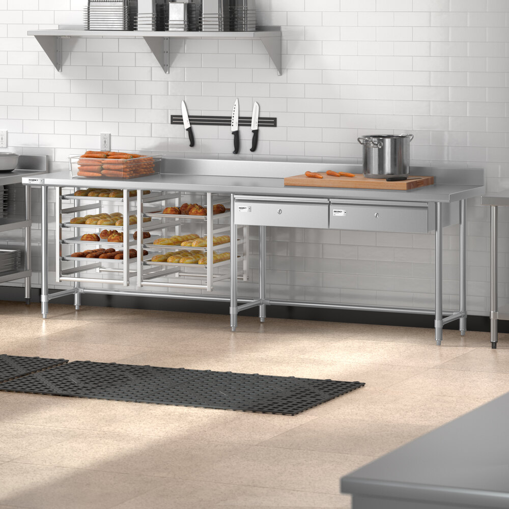 A stainless steel commercial kitchen work table with an open base, multiple shelves holding trays of baked goods, a backsplash, and drawers, set against a white tiled wall.