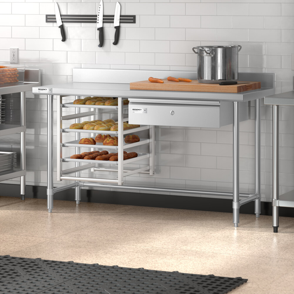 A stainless steel work table with an open base, a drawer, a cutting board with carrots, a large pot, and multiple trays of baked goods underneath, set in a commercial kitchen.