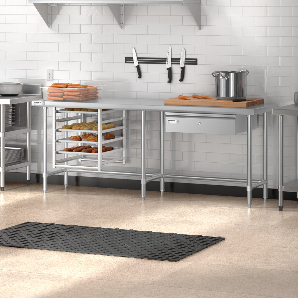 A stainless steel commercial kitchen work table with an open base, a rack holding trays of baked goods, a cutting board with knives and a pot on top, and a wall-mounted magnetic knife strip above.
