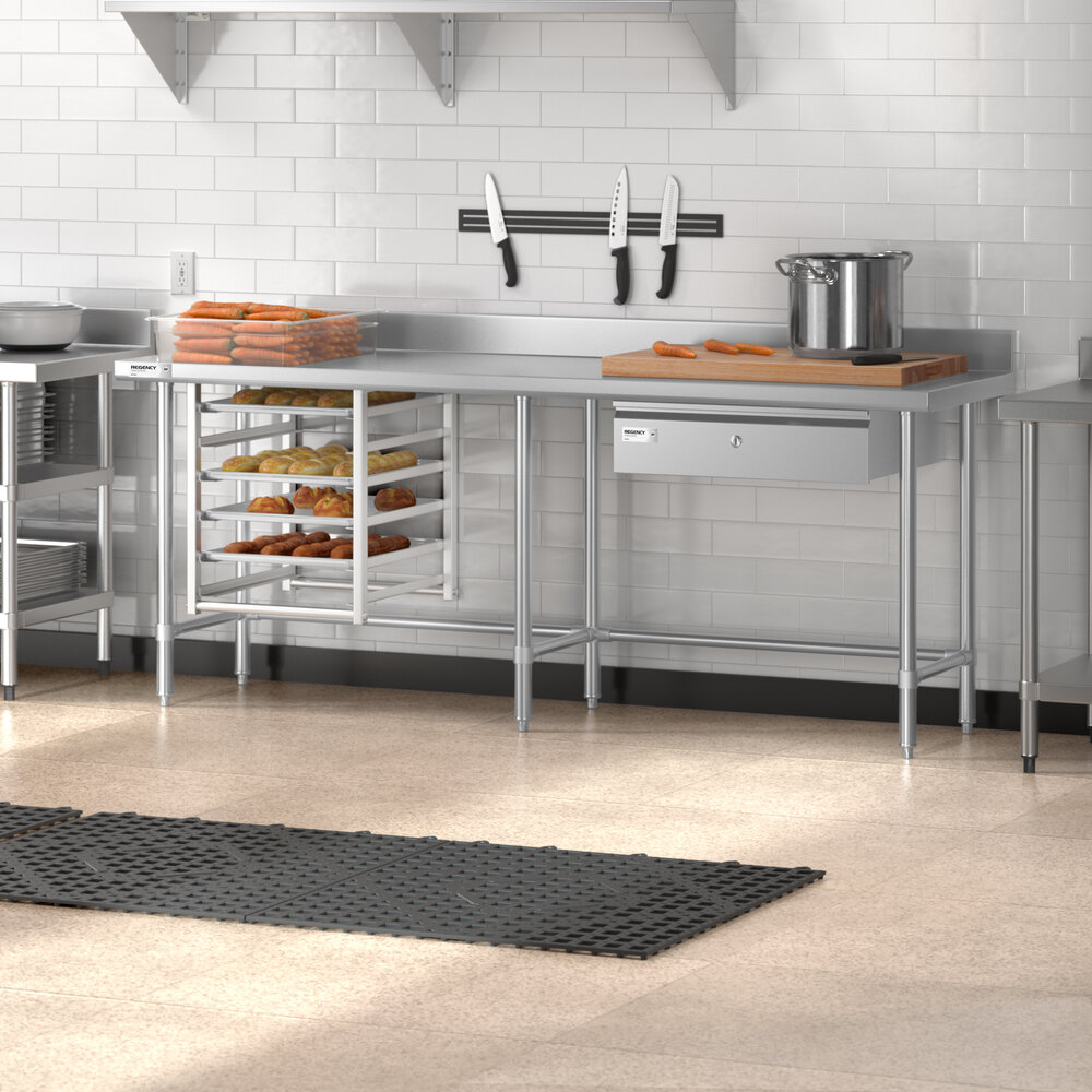 A stainless steel work table with an open base, a 4-inch backsplash, and a drawer, shown in a commercial kitchen setting with baked goods, knives, a pot, and a cutting board on top.