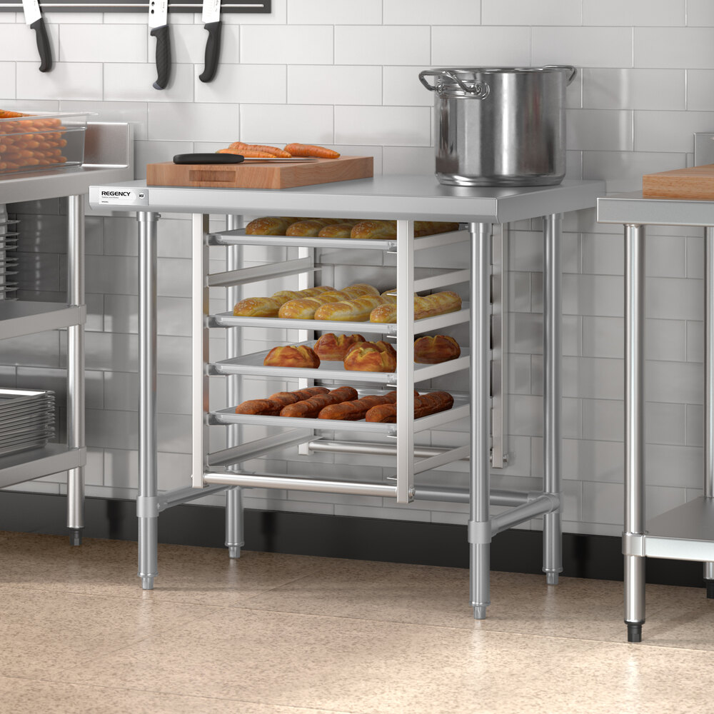 A stainless steel work table with an open base, featuring multiple racks holding trays of baked goods in a commercial kitchen setting.