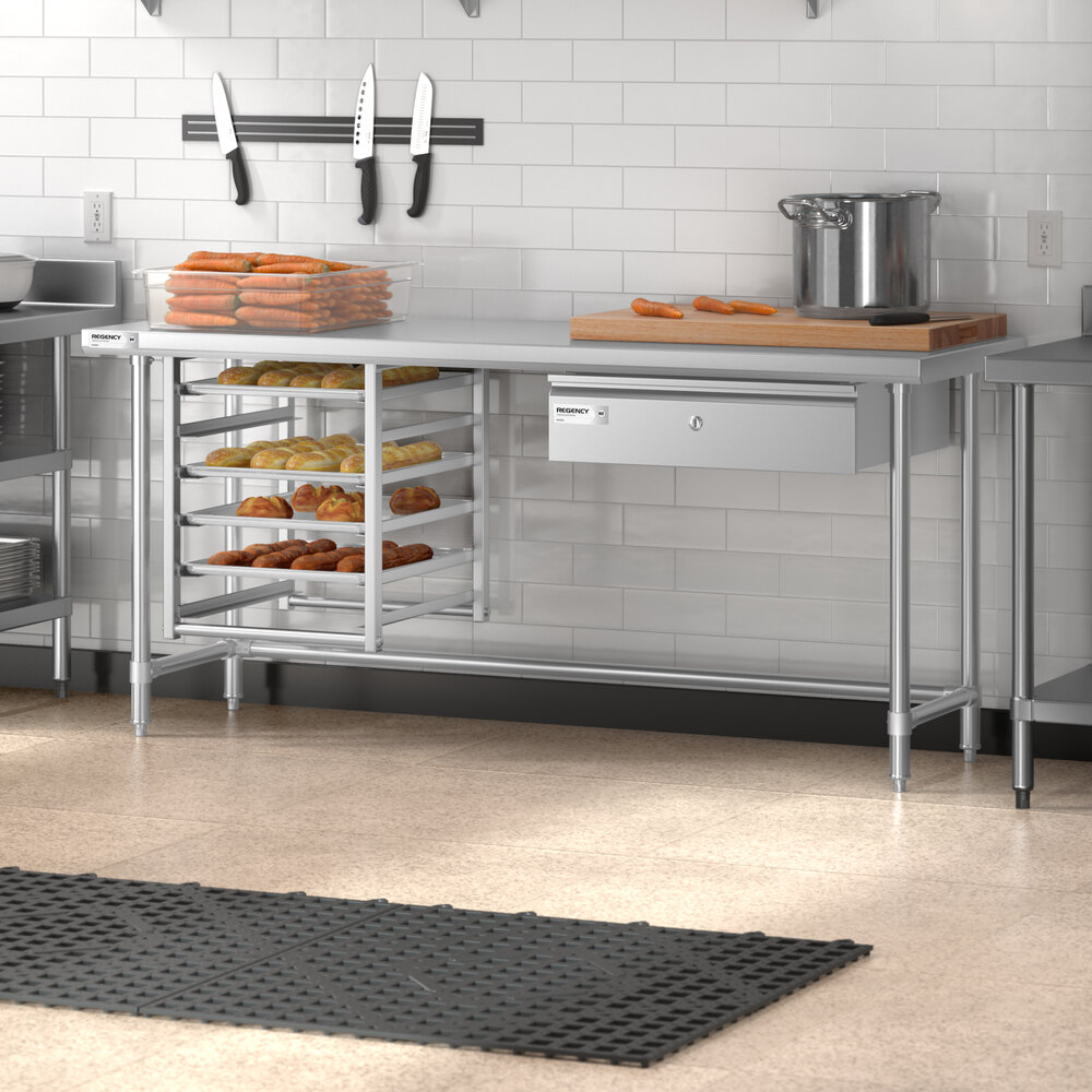 A stainless steel work table with an open base, featuring multiple shelves holding trays of baked goods, a drawer, and a countertop with kitchen utensils and food items in a commercial kitchen setting.