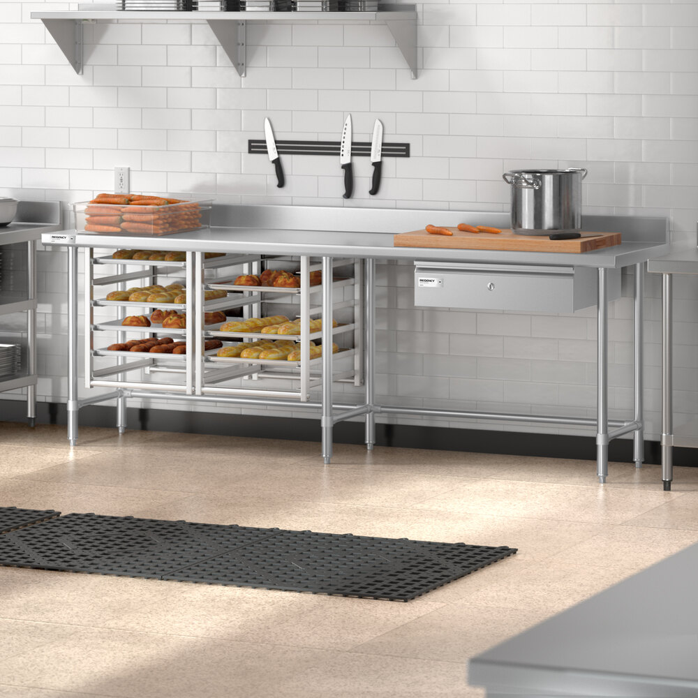 A stainless steel commercial kitchen work table with an open base, multiple shelves holding trays of baked goods, a 4-inch backsplash, and various kitchen items on top including a pot, cutting boards, and knives mounted on the wall.