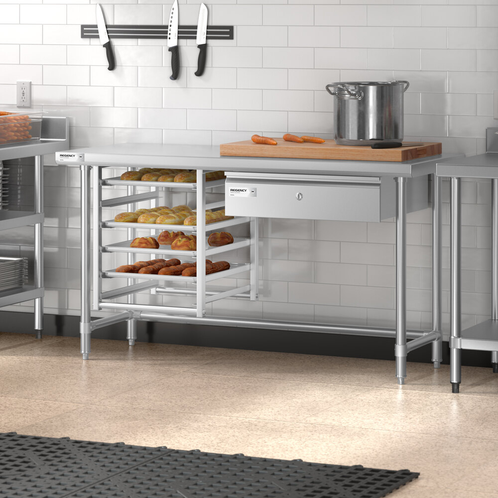 A stainless steel work table with an open base, multiple shelves holding baked goods, a drawer, and a cutting board with carrots and a pot on top, set in a commercial kitchen.