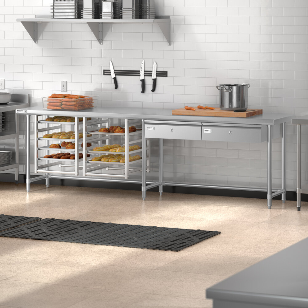 A stainless steel commercial kitchen work table with open shelving, drawers, and various kitchen items including trays of baked goods, a pot, and knives mounted on the wall.