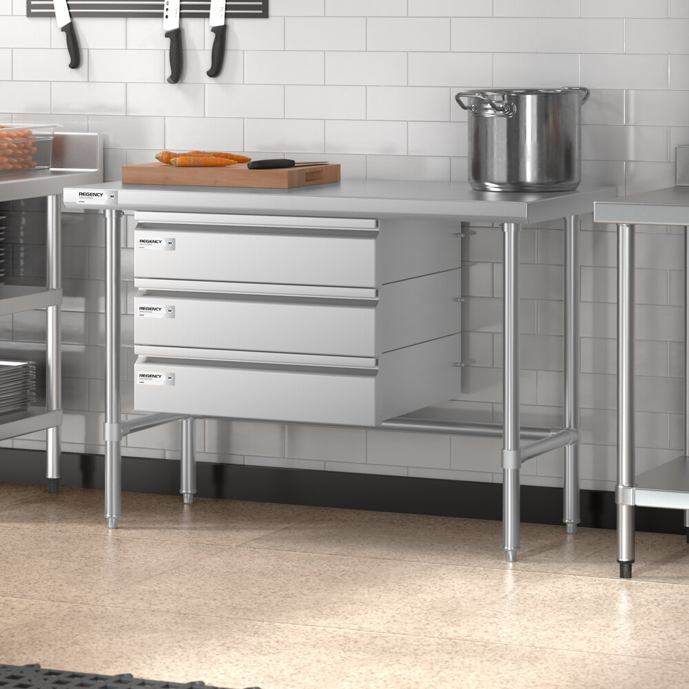 A stainless steel work table with three drawers, an open base, and a flat top surface, situated in a commercial kitchen setting.
