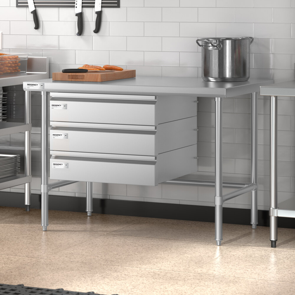 A stainless steel work table with three drawers, an open base, and a pot and cutting board on top, situated in a commercial kitchen setting.