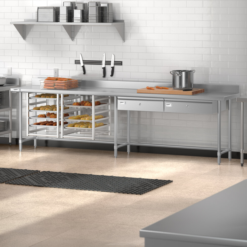 A stainless steel commercial kitchen work table with an open base, two drawers, a 4-inch backsplash, and racks holding trays of baked goods underneath.