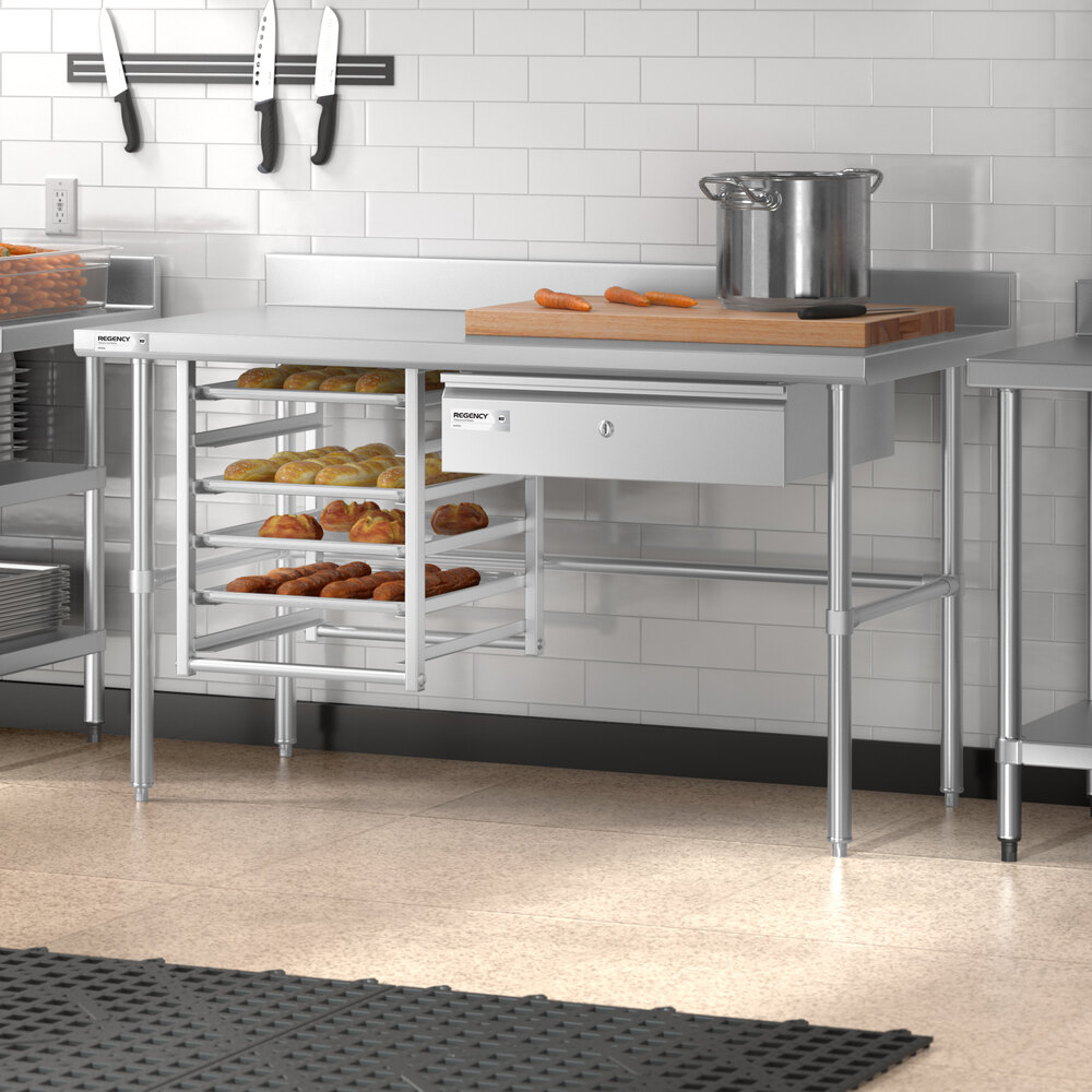 A stainless steel work table with an open base, a drawer, a rack holding trays of baked goods, a cutting board with carrots, and a pot on top, set in a commercial kitchen.
