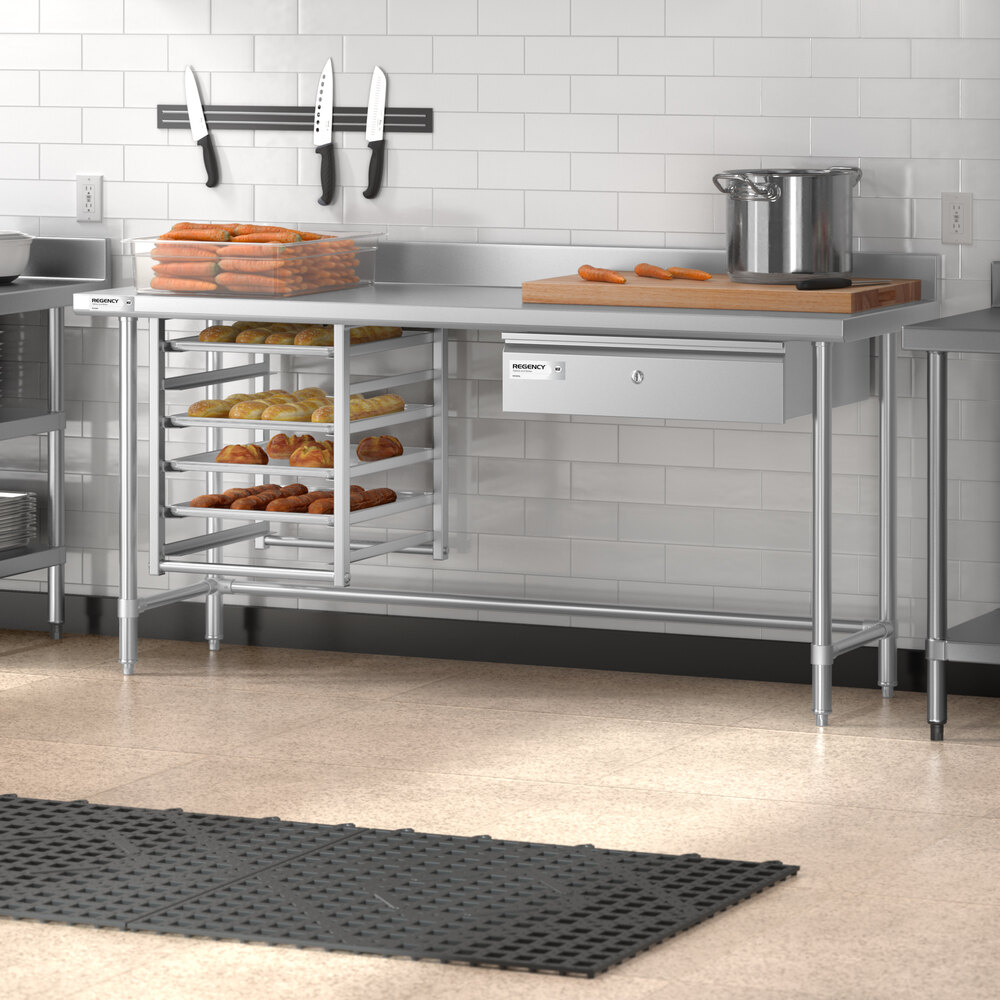 A stainless steel work table with an open base, a 4-inch backsplash, a mounted drawer, and multiple trays of baked goods underneath, set in a commercial kitchen environment.